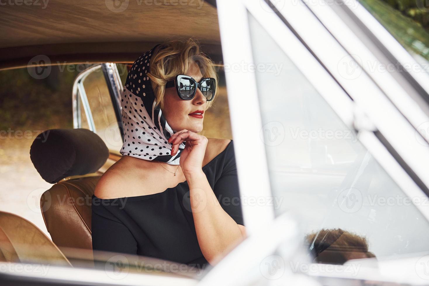 mulher loira de óculos escuros e vestido preto senta-se no velho carro clássico vintage foto