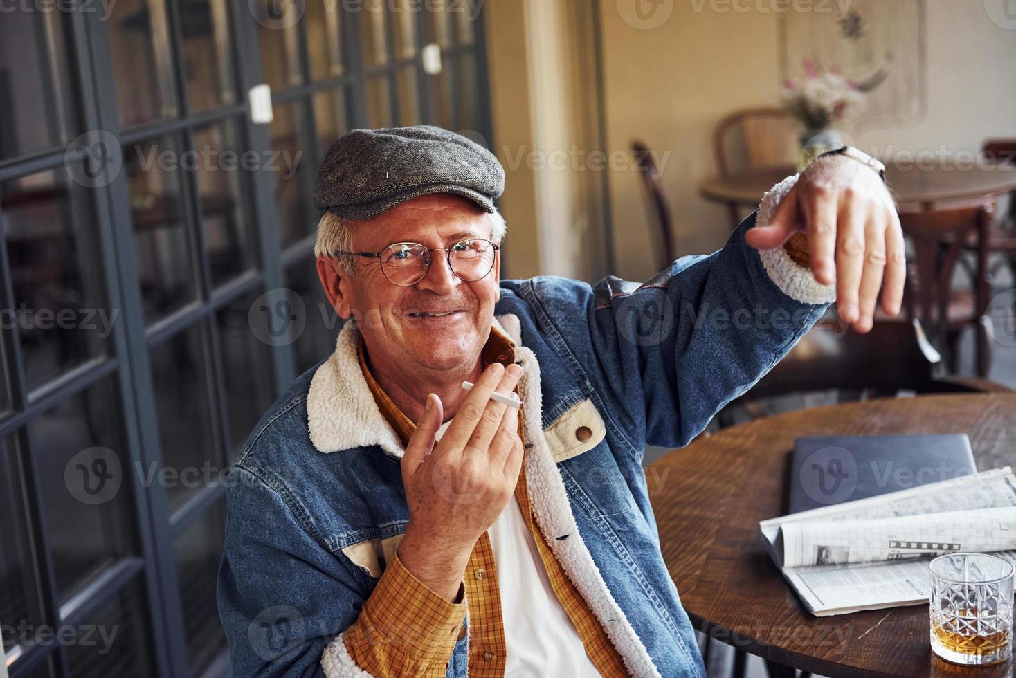 elegante sênior em roupas da moda e de óculos senta-se no café com cigarro e fala foto