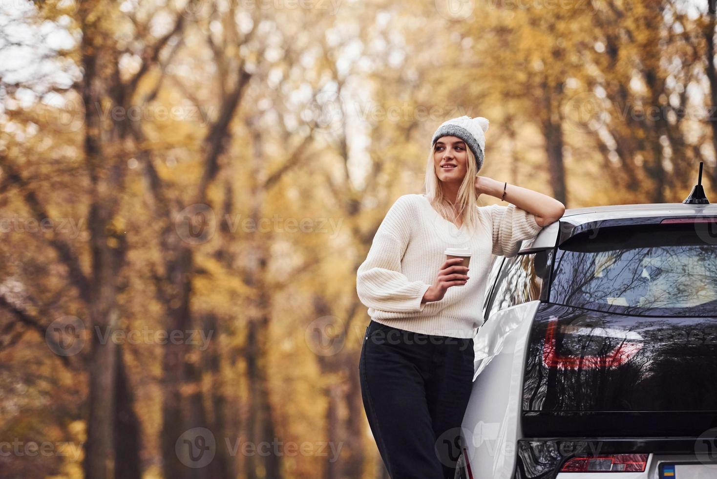 com copo de bebida nas mãos. menina tem viagem de outono de carro. automóvel novo e moderno na floresta foto