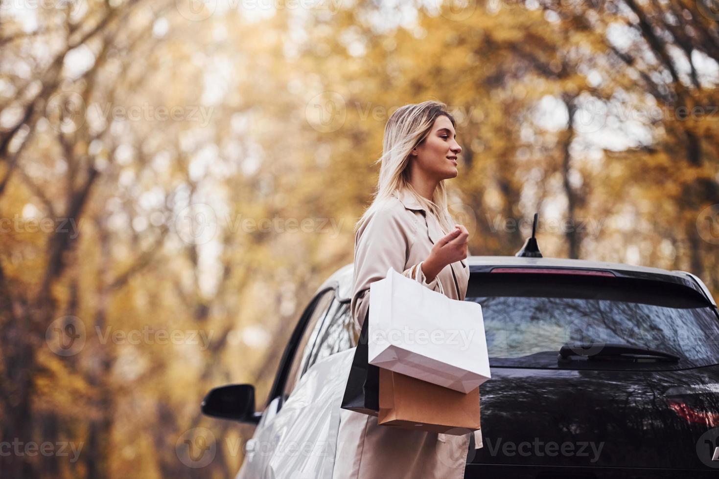 menina caminha perto do carro com sacolas de compras nas mãos. automóvel novo e moderno na floresta foto