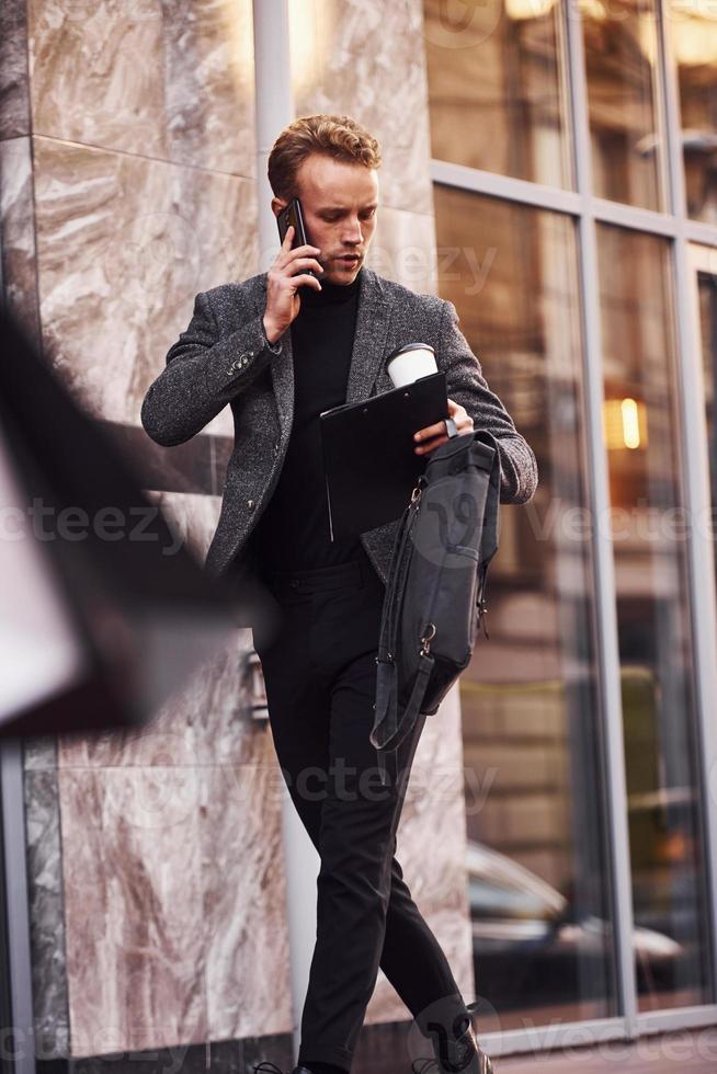 homem com roupa formal elegante com copo de bebida e bloco de notas nas mãos está do lado de fora contra o edifício moderno conversando ao telefone foto