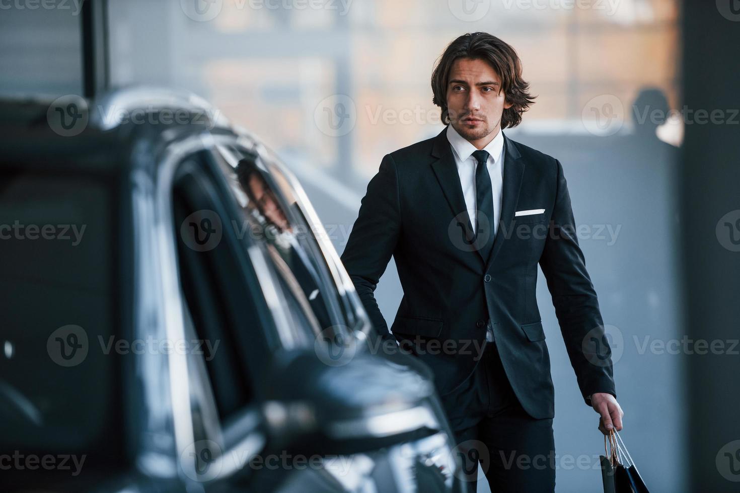 retrato do jovem empresário bonito de terno preto e gravata e com sacola de compras perto do carro moderno foto