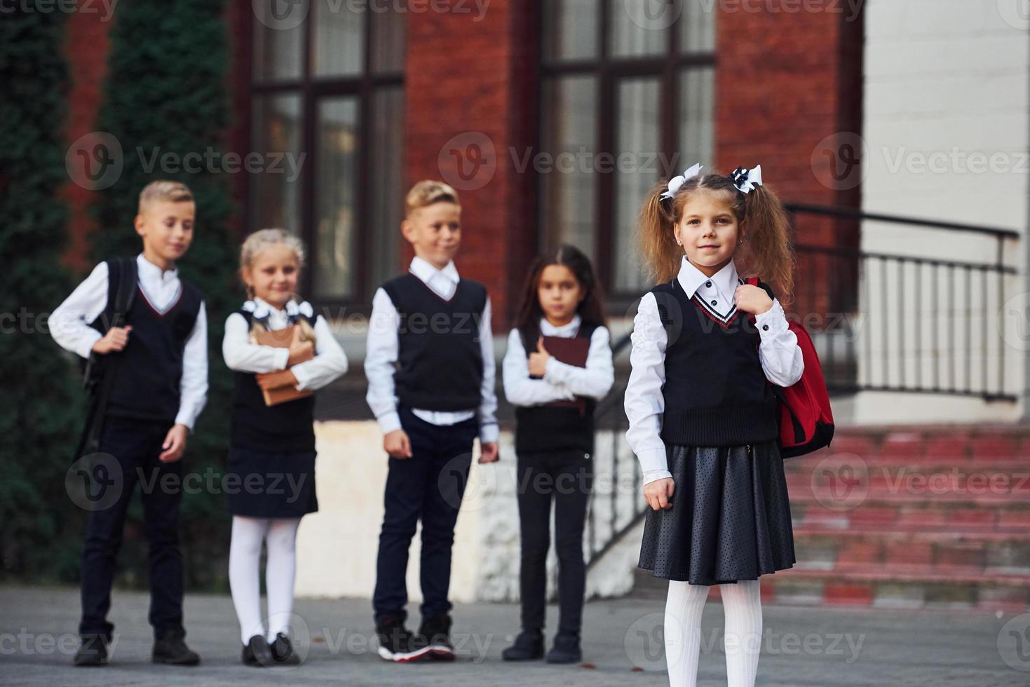 grupo de crianças em uniforme escolar posando para a câmera ao ar livre juntos perto do prédio da educação foto