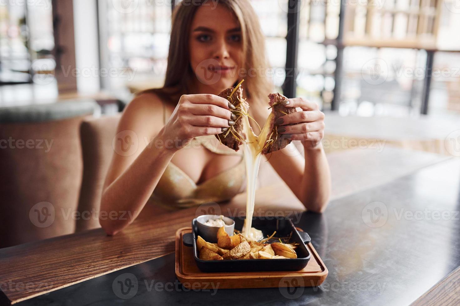 mulher loira sexy com roupa quente senta-se à mesa e come fast-food dentro de casa no café foto