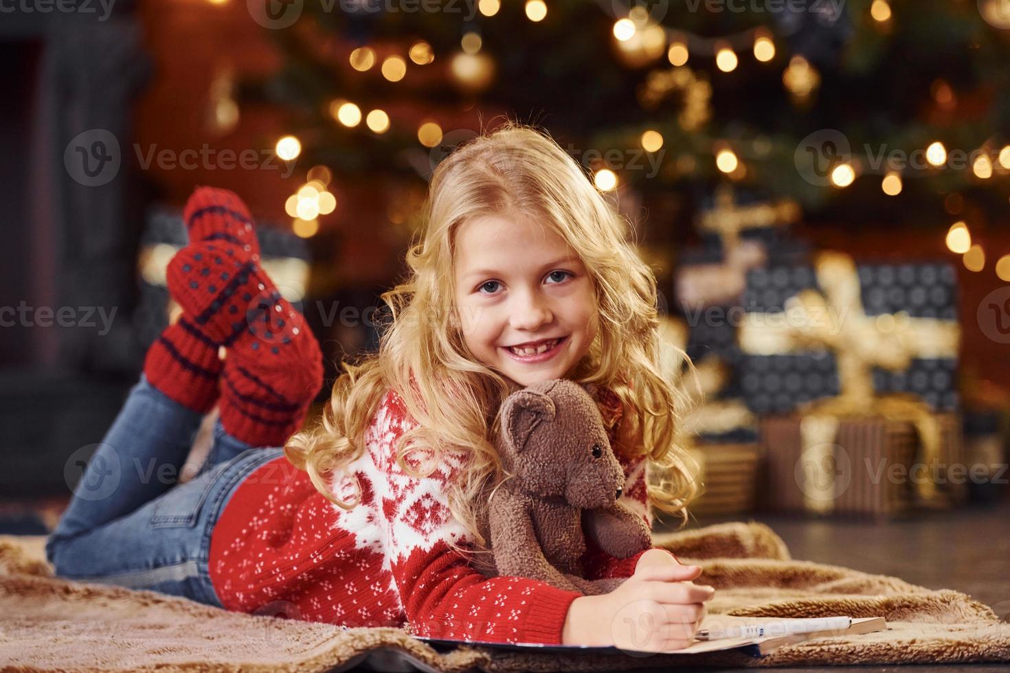 menina bonitinha no suéter festivo vermelho deitado com ursinho de pelúcia dentro de casa comemorando o ano novo foto