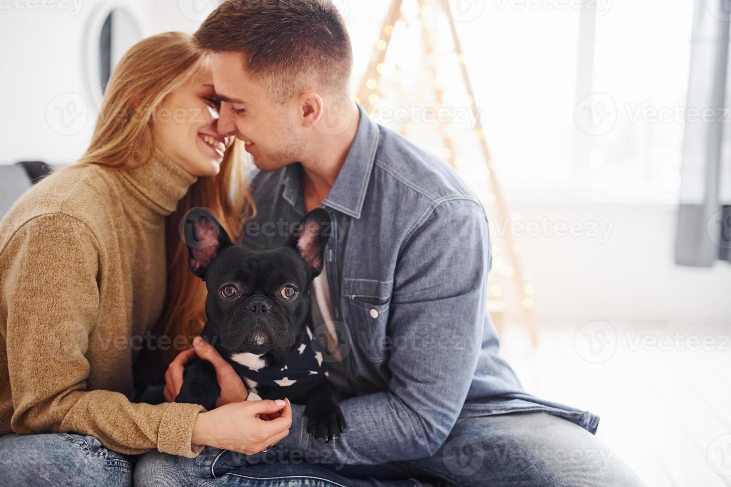 feliz jovem casal moderno sentado no sofá em casa com a árvore de natal com seu cachorro fofo foto