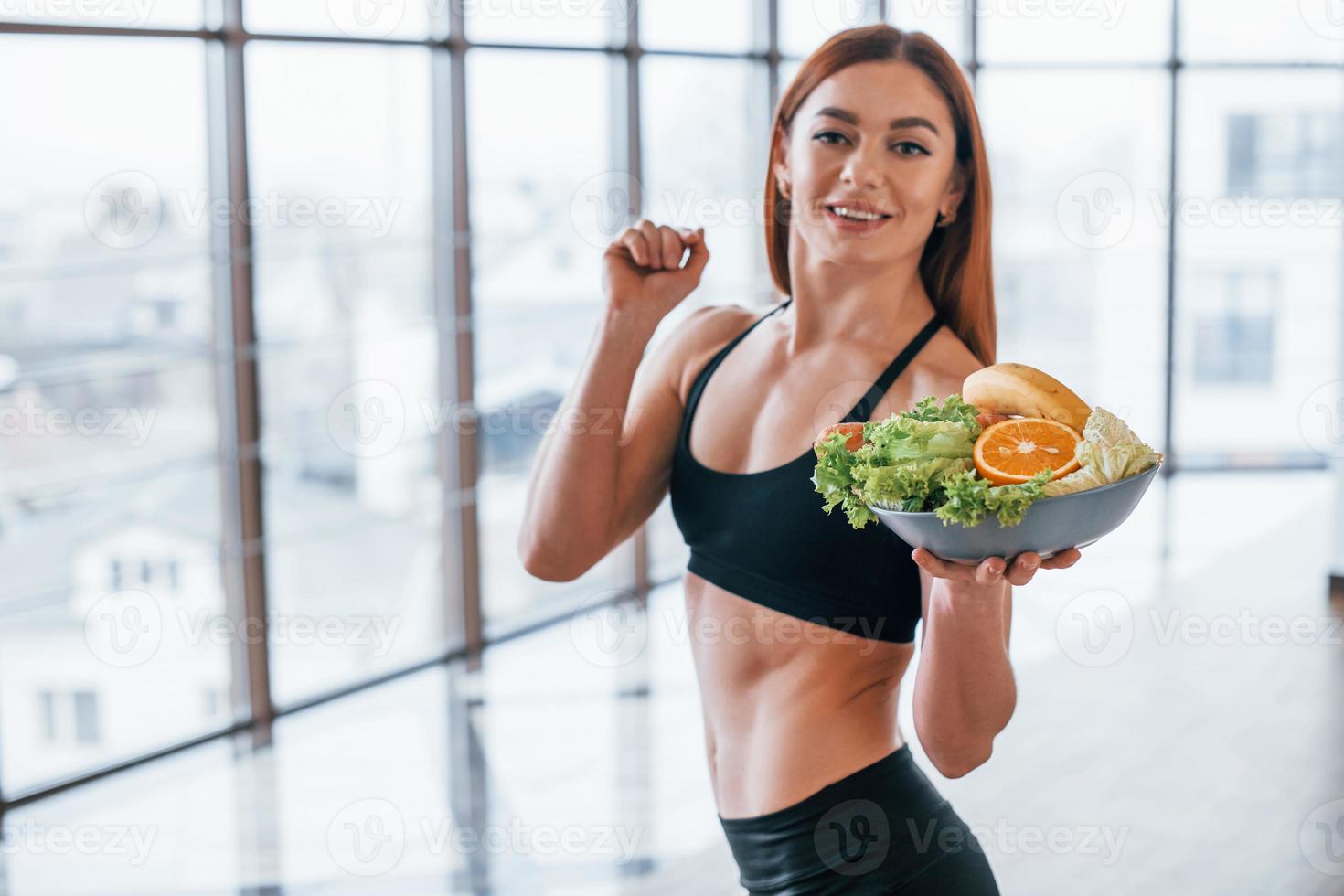 alegre mulher esportiva fica dentro de casa com comida saudável nas mãos foto