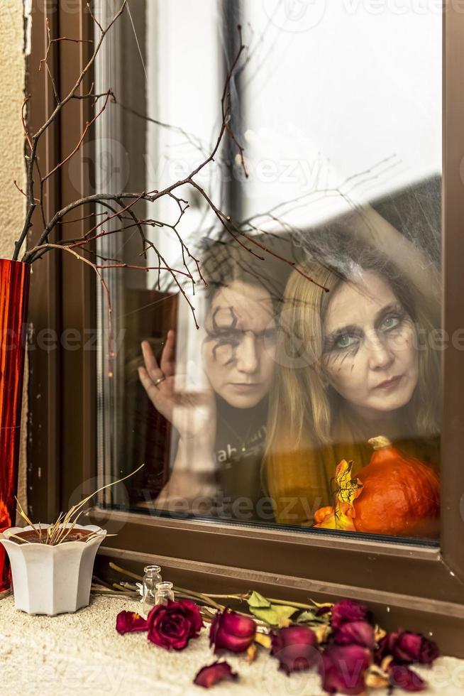 duas bruxas olham através de uma teia de aranha ameaçadoramente pela janela contra o pano de fundo das decorações de halloween. baile de máscaras, festa de halloween foto