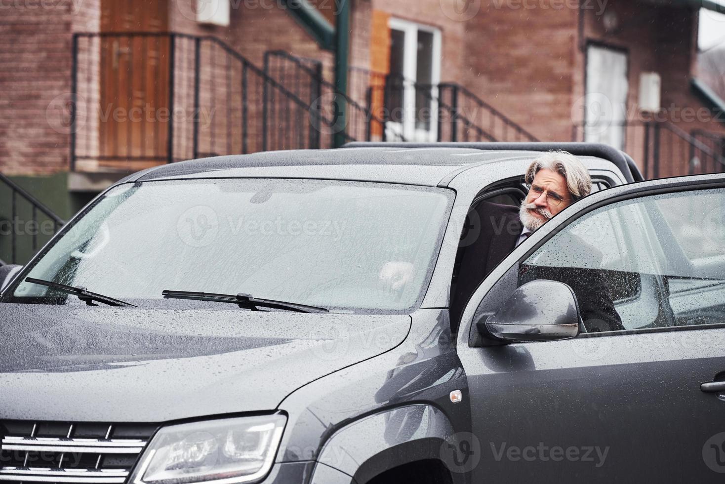 elegante homem sênior com cabelos grisalhos e barba está ao ar livre na rua sentado em seu carro foto