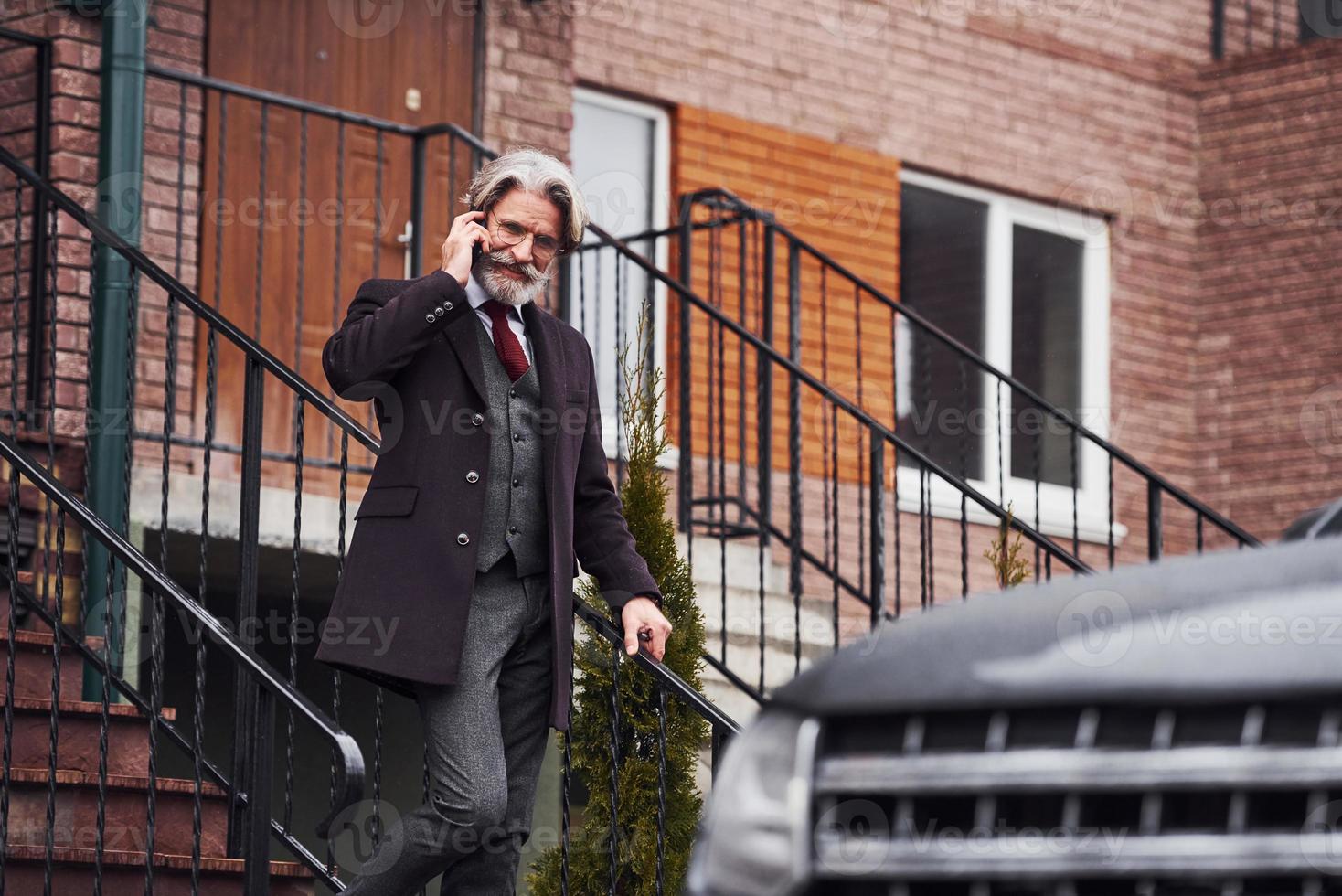 elegante homem sênior com cabelos grisalhos e barba está ao ar livre na rua perto de seu carro foto