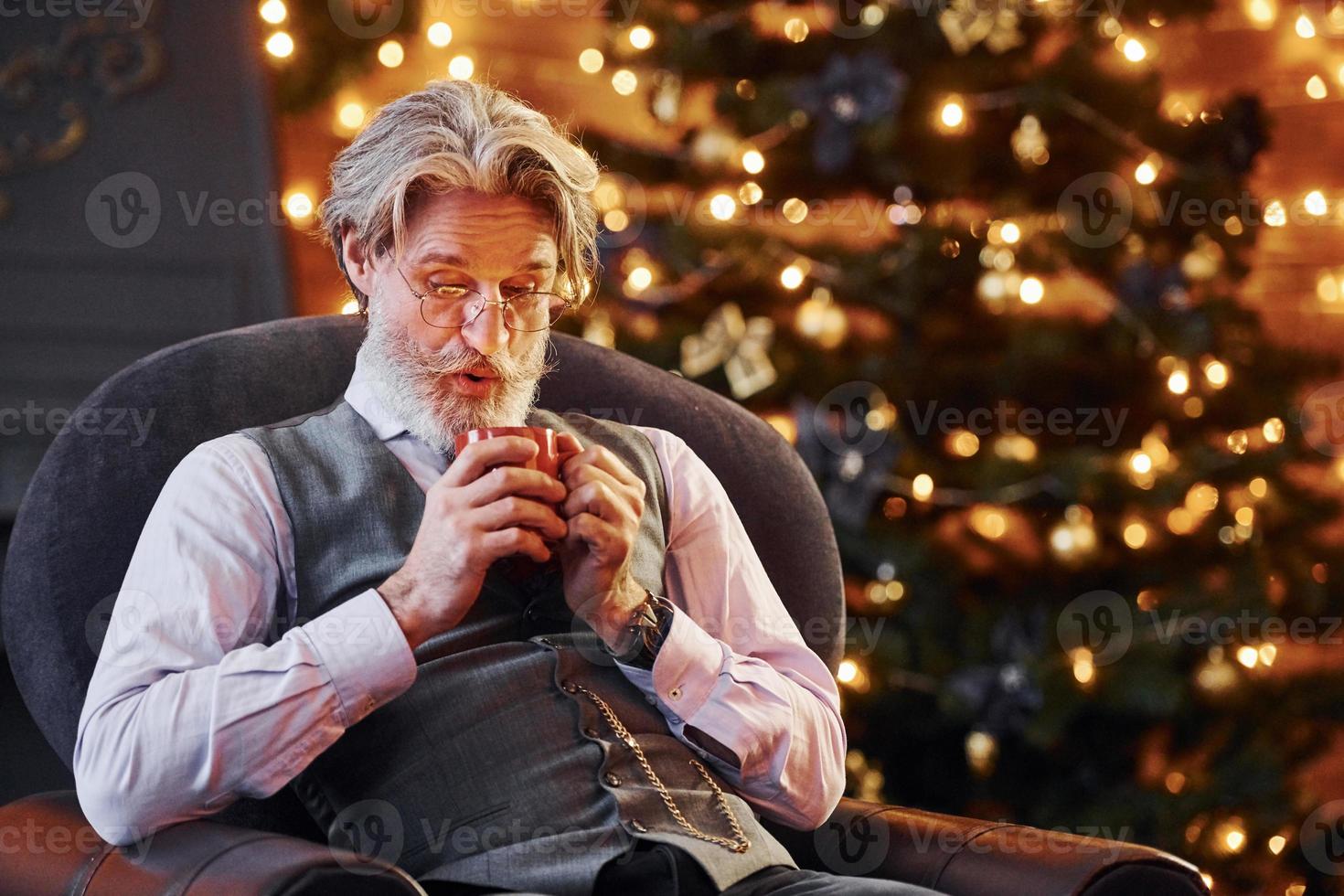 retrato de elegante sênior com cabelos grisalhos e barba está na sala de natal decorada com copo de bebida foto