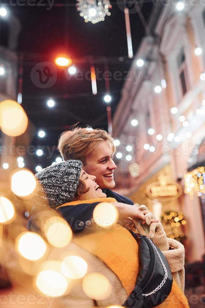 casal jovem positivo em roupas quentes, abraçando-se na rua decorada de natal foto