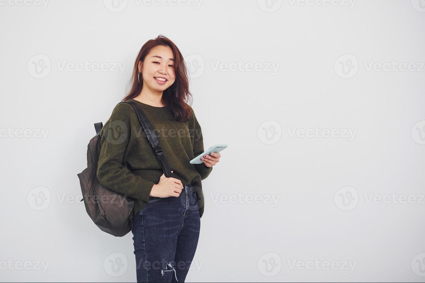 retrato de uma jovem asiática feliz com mochila e telefone que está dentro de casa no estúdio contra um fundo branco foto