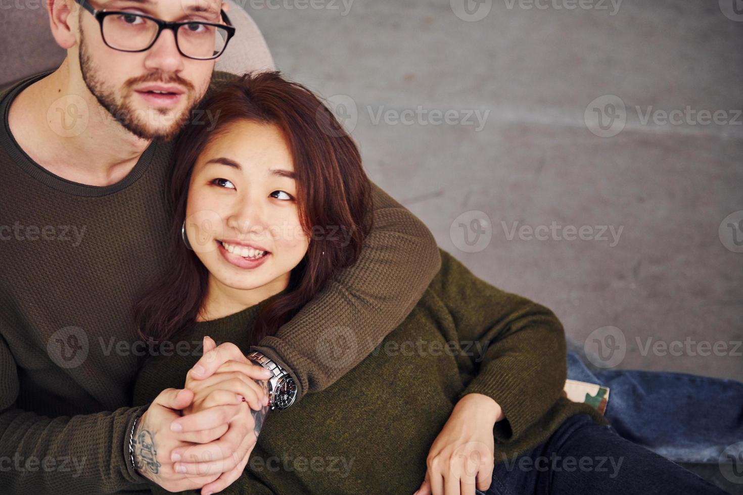 feliz casal multiétnico em roupas casuais sentados juntos dentro de casa no estúdio. cara caucasiano com namorada asiática foto