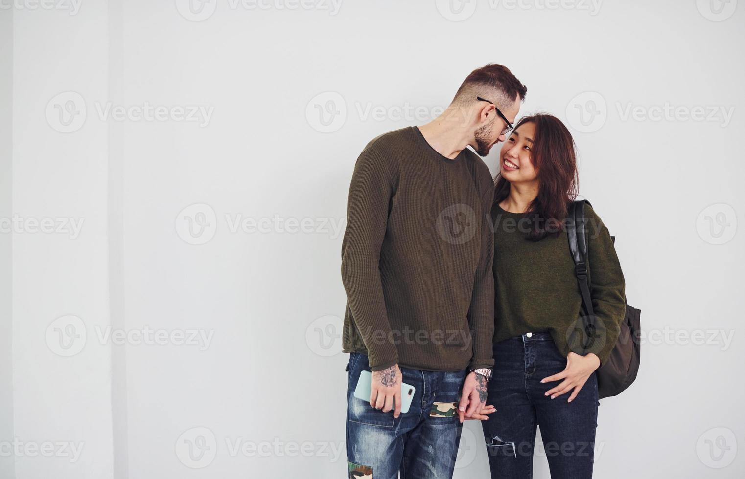 alegre casal multiétnico com mochila e telefone juntos dentro de casa no estúdio contra fundo branco foto