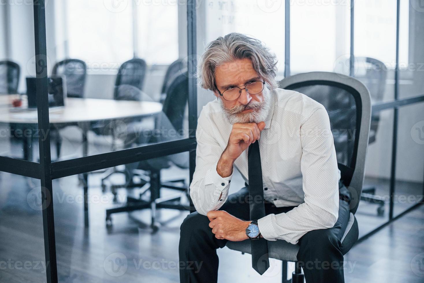 empresário maduro sério com cabelos grisalhos e barba em roupas formais, sentado no escritório foto