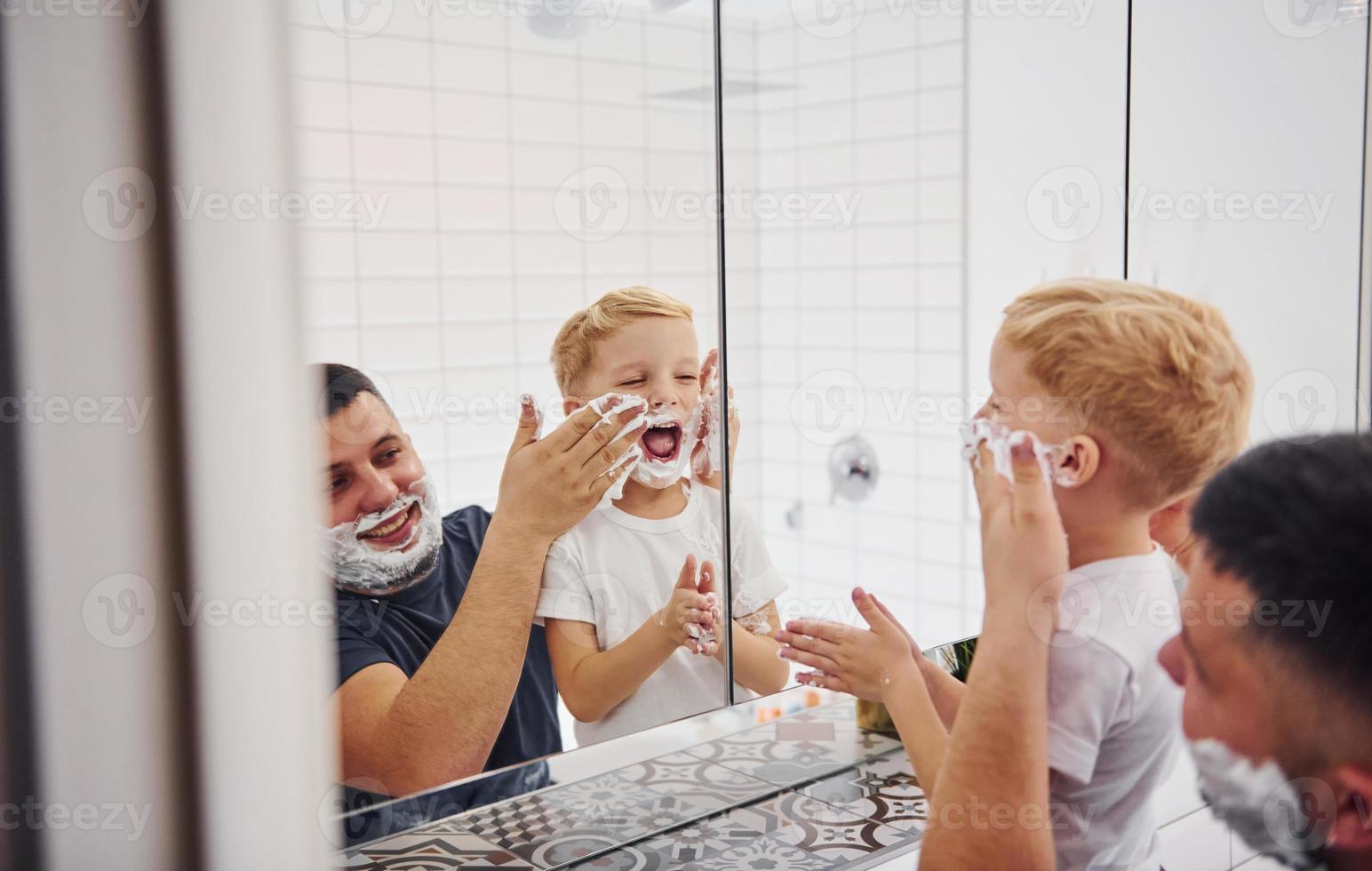 pai com seu filho está no banheiro se divertindo usando gel de barbear e olhando no espelho foto