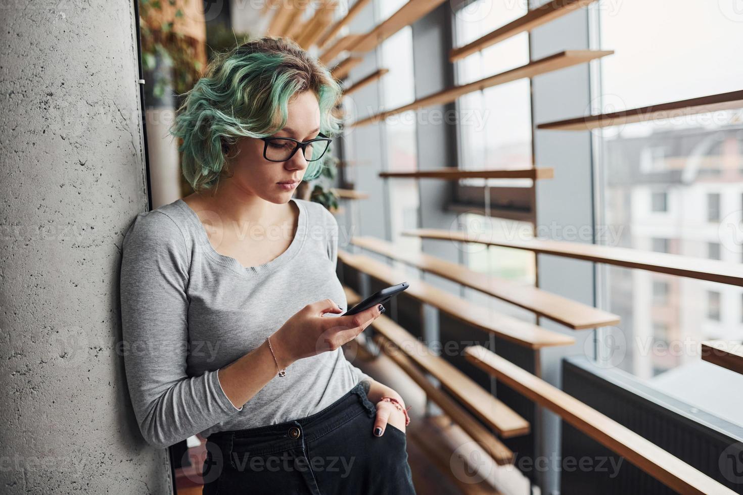 garota alternativa em roupas casuais e com cabelo verde em pé dentro de casa durante o dia com o telefone na mão foto