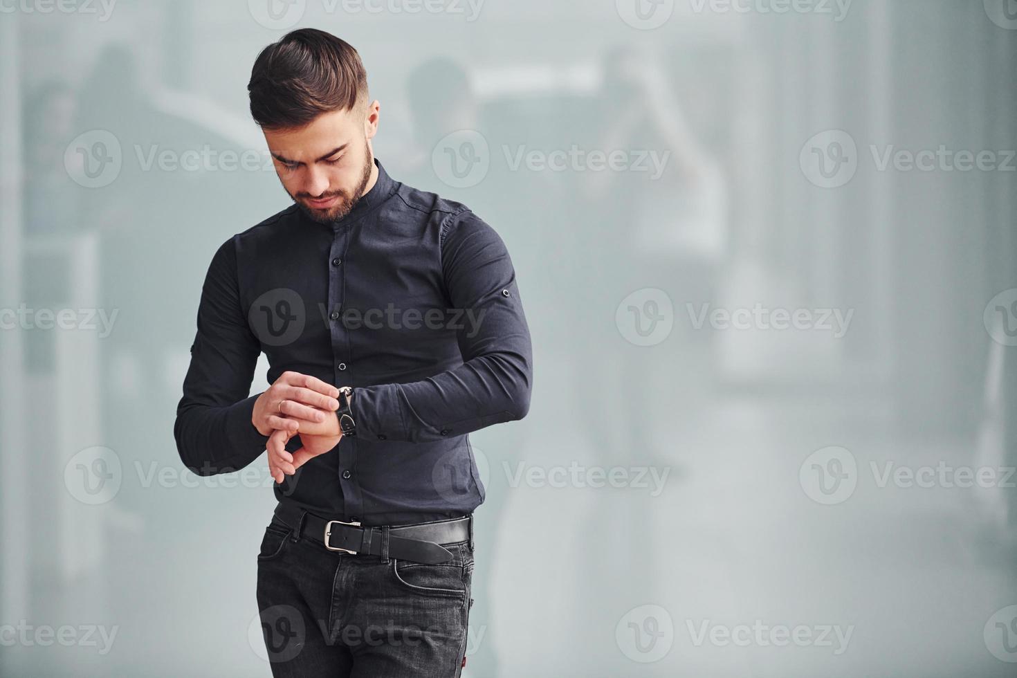 jovem barbudo em roupas elegantes em pé dentro de casa contra um fundo cinza foto