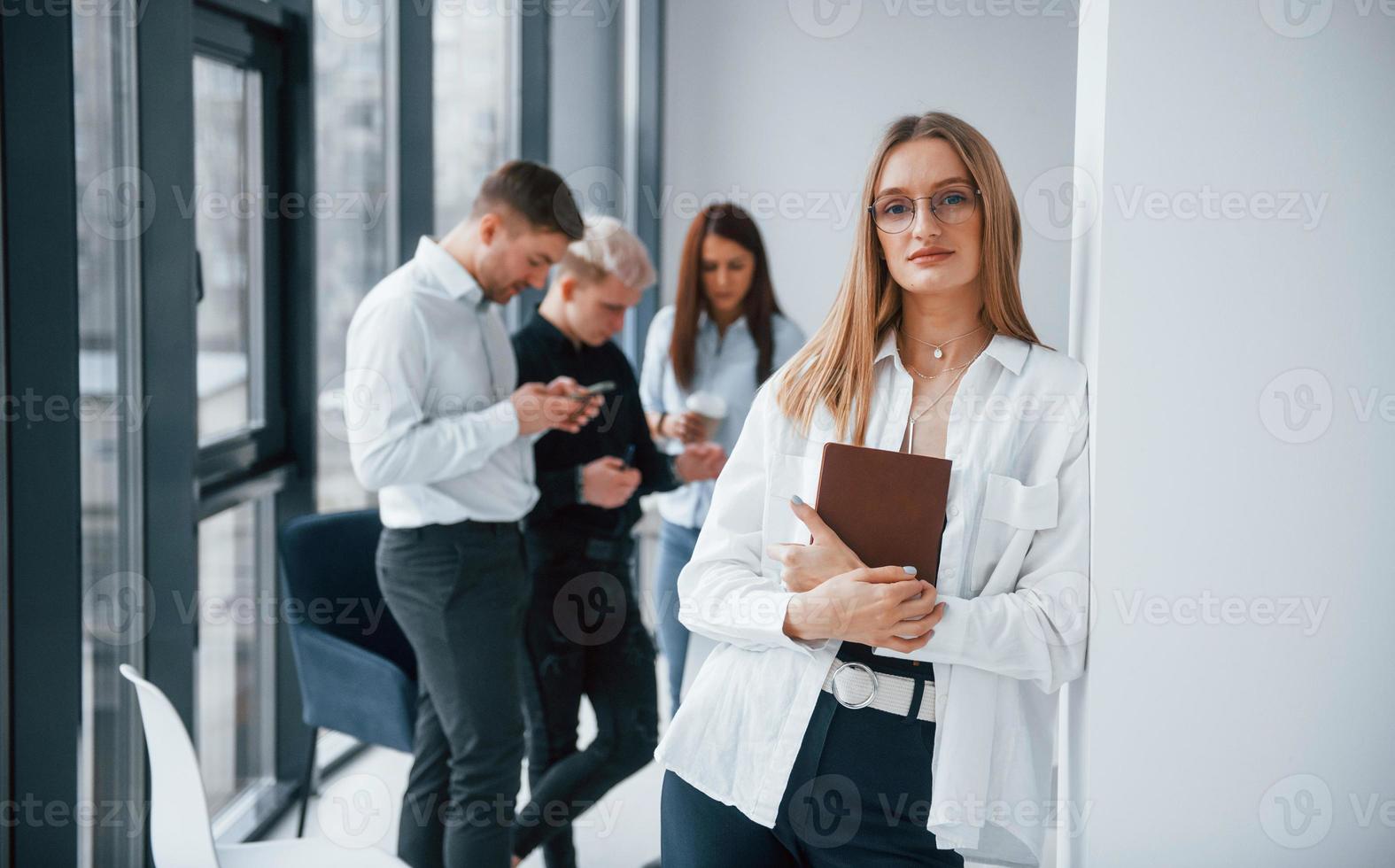 retrato de uma loira alegre com um bloco de notas que fica na frente de uma jovem equipe de sucesso que trabalha e se comunica dentro de casa no escritório foto
