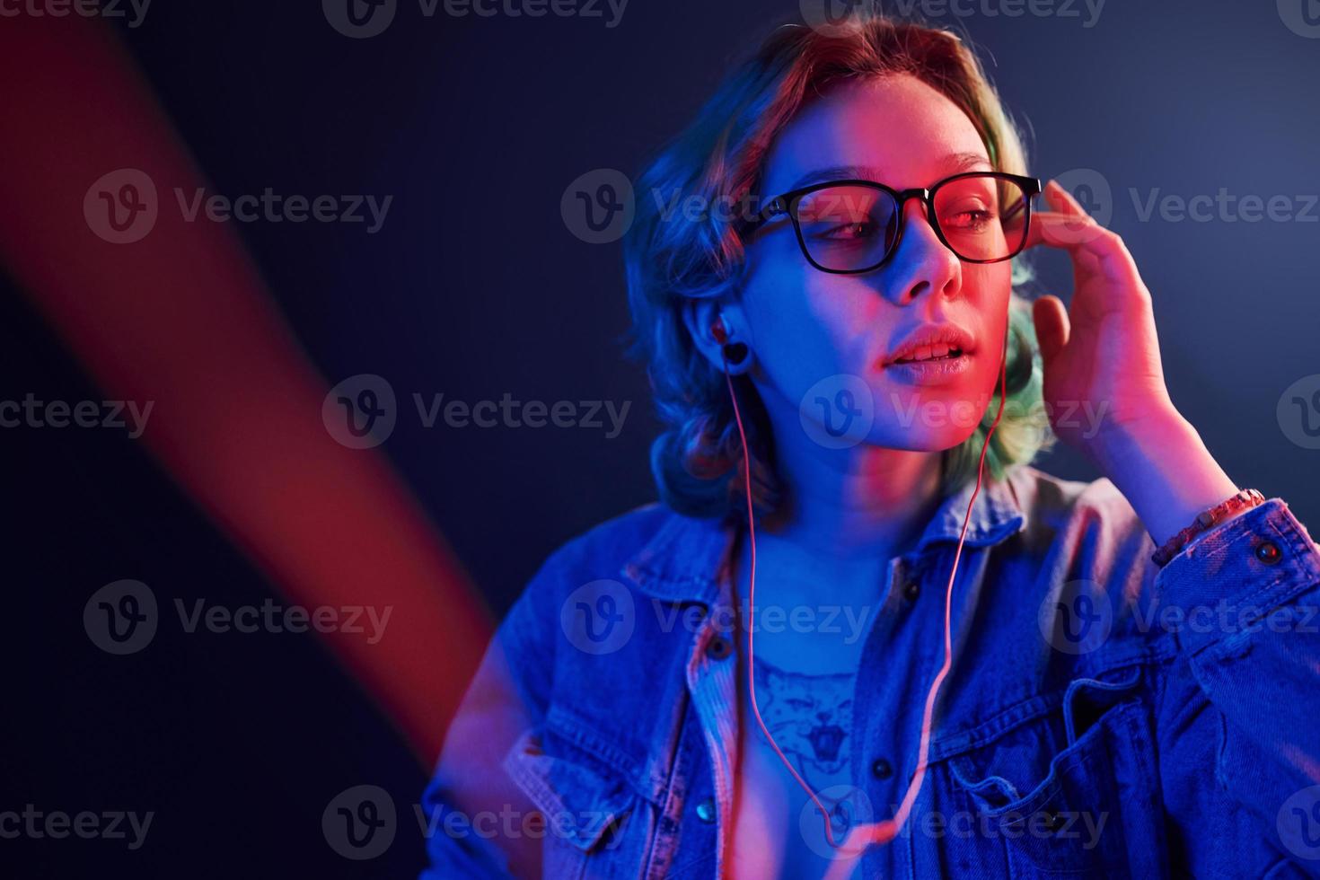 retrato de jovem alternativa em óculos com cabelo verde em luz neon vermelha e azul no estúdio que ouve música em fones de ouvido foto