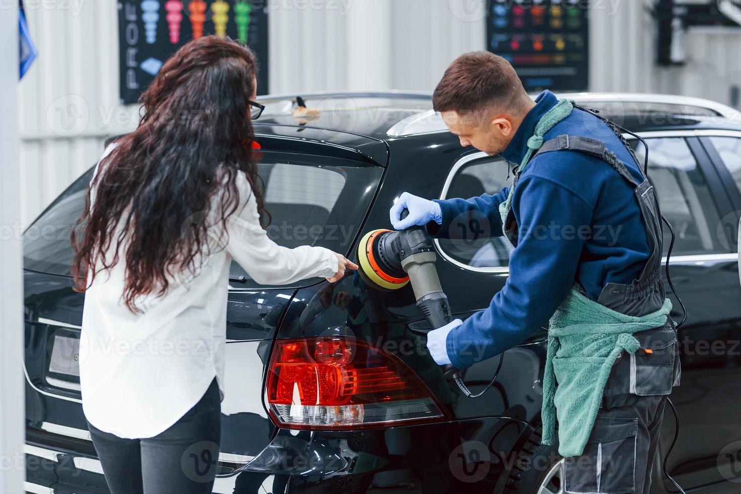 cliente do sexo feminino falando o que fazer. trabalhador masculino de uniforme limpando carro novo e moderno dentro de casa. concepção de serviço foto