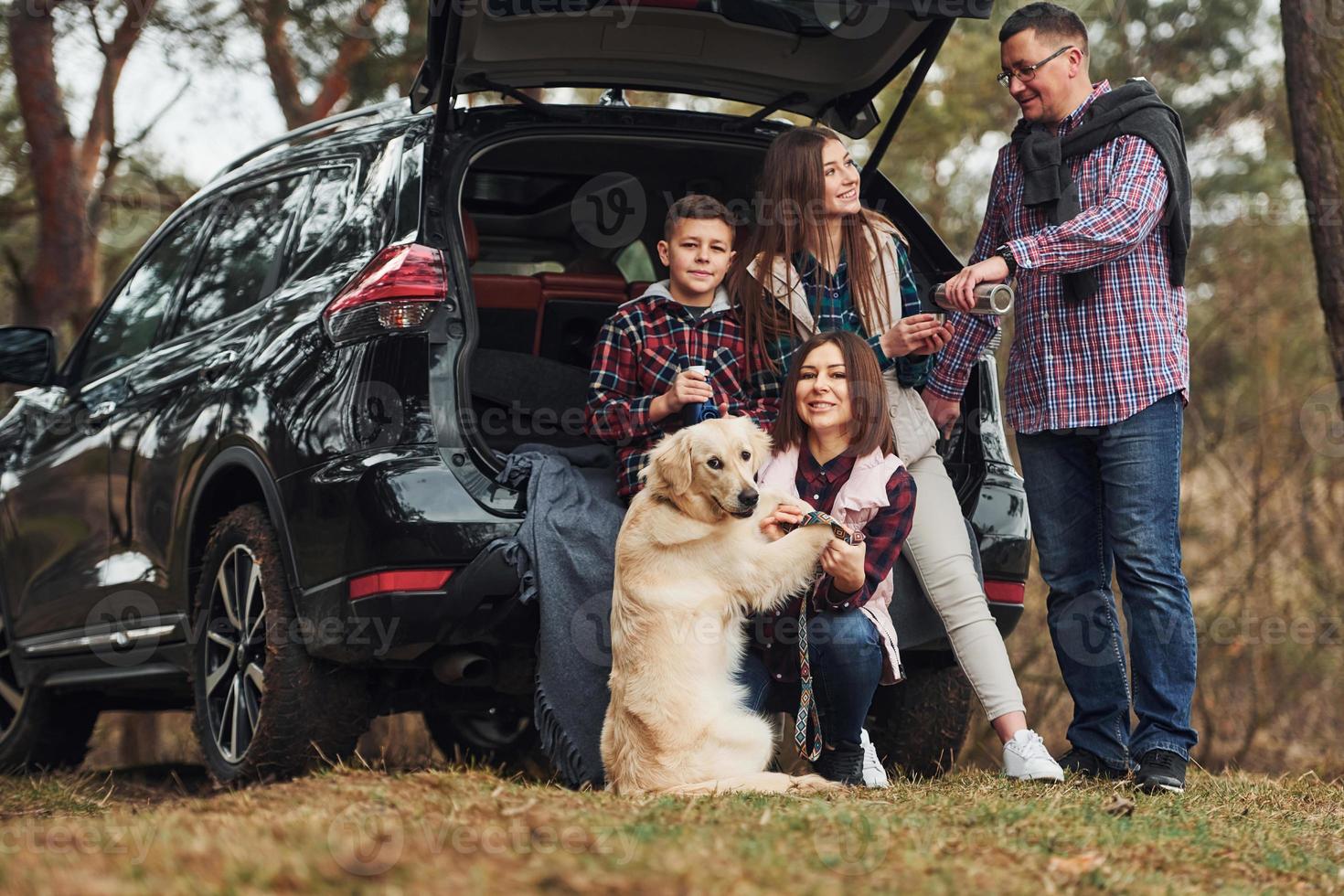 família feliz se diverte com seu cachorro perto de carro moderno ao ar livre na floresta foto