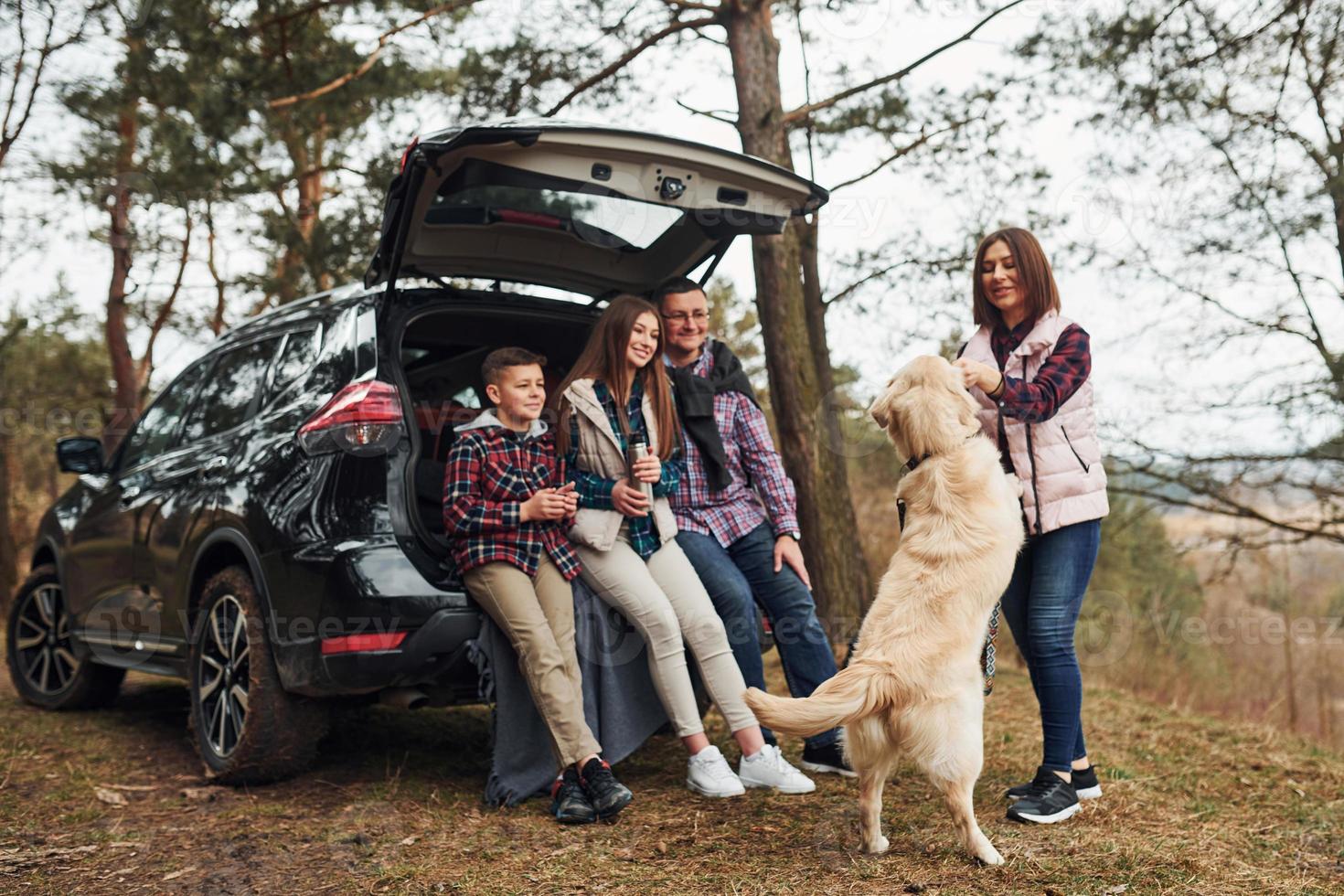 família feliz se diverte com seu cachorro perto de carro moderno ao ar livre na floresta foto