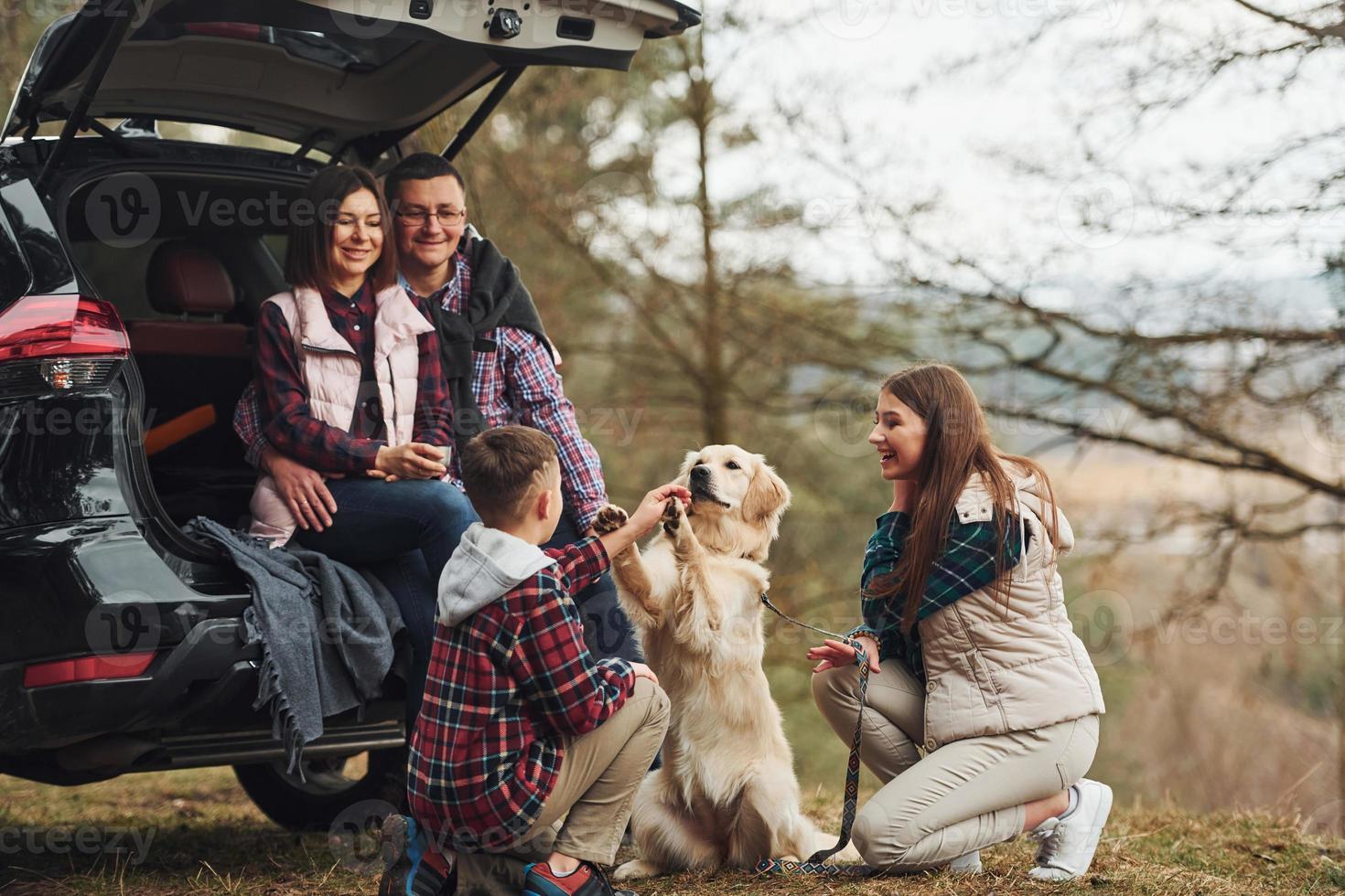 família feliz se diverte com seu cachorro perto de carro moderno ao ar livre na floresta foto