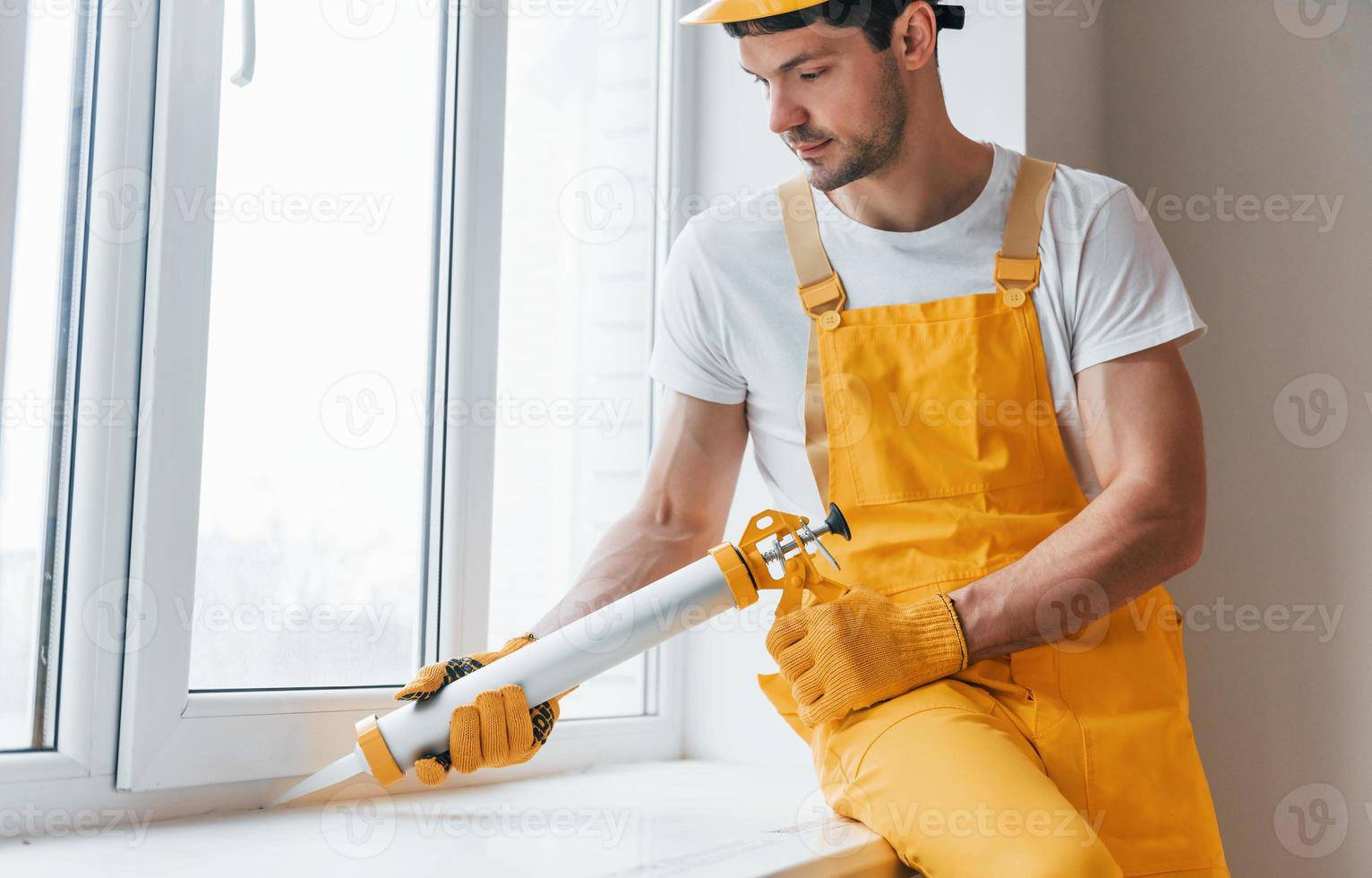 faz-tudo de uniforme amarelo trabalha com cola para janela dentro de casa. concepção de renovação de casa foto