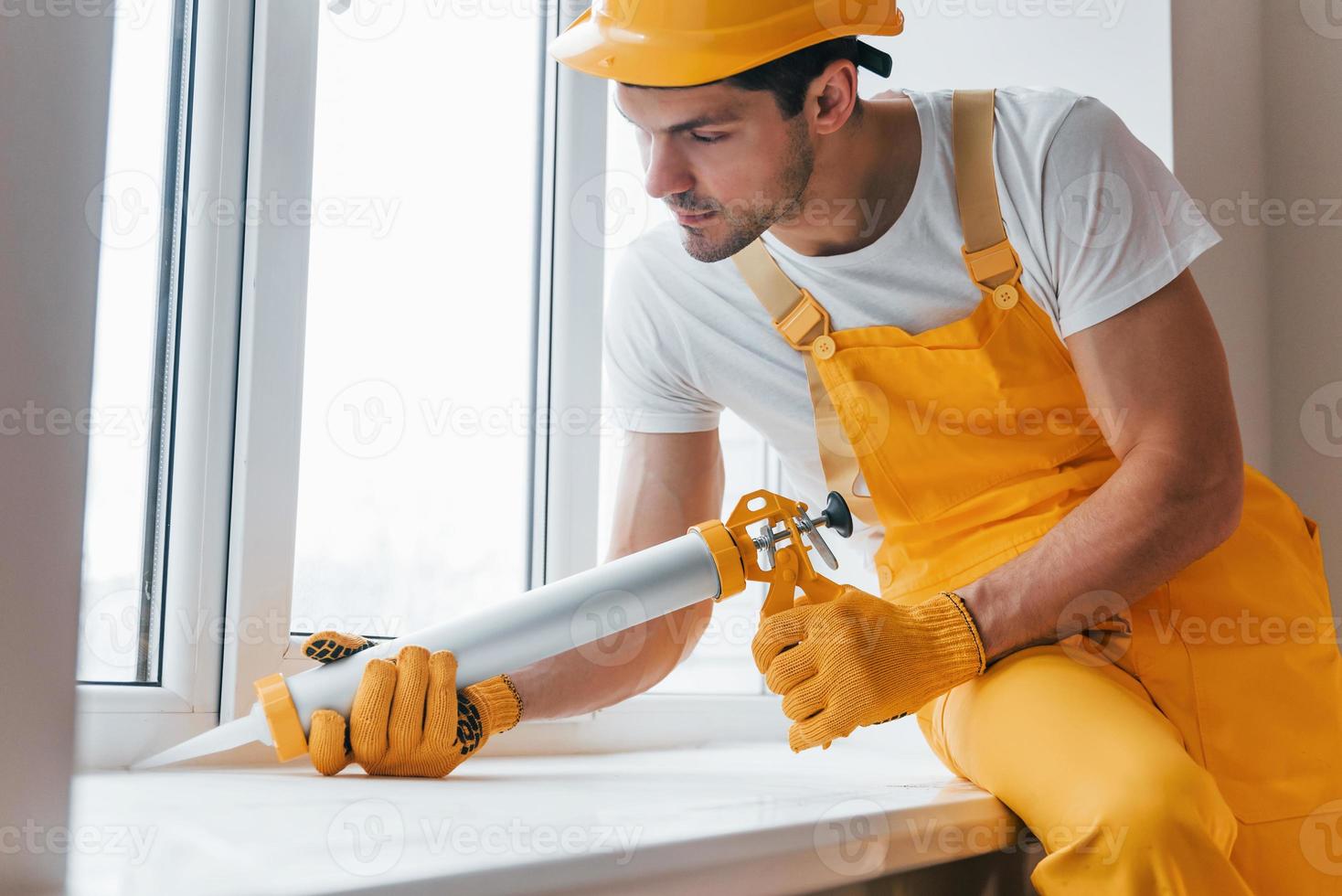 faz-tudo de uniforme amarelo trabalha com cola para janela dentro de casa. concepção de renovação de casa foto