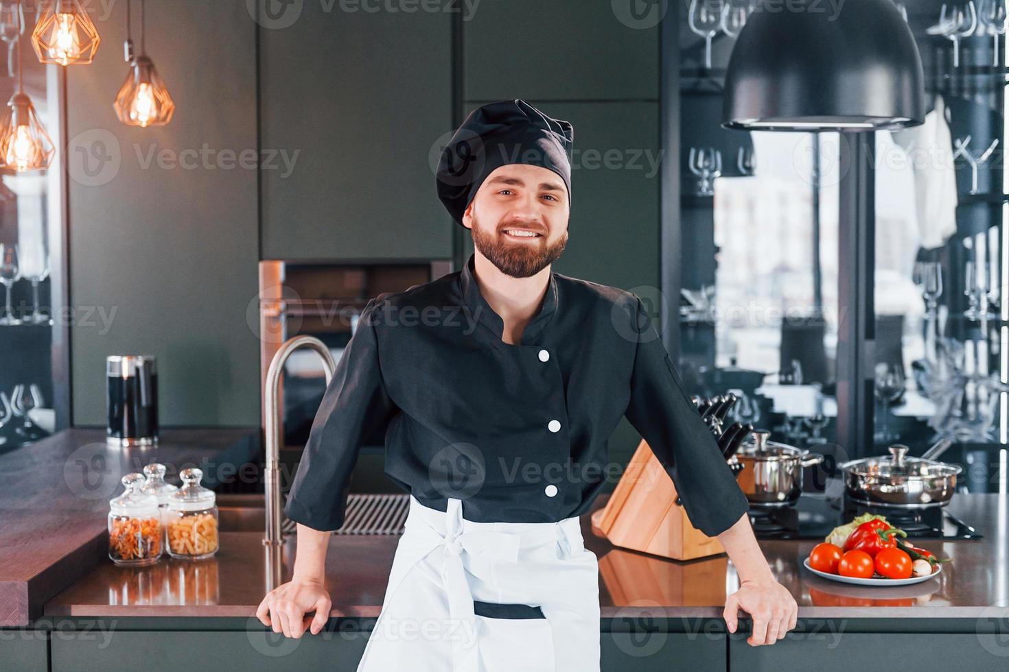 cozinheiro profissional jovem chef de uniforme em pé perto da mesa e posando para uma câmera na cozinha foto