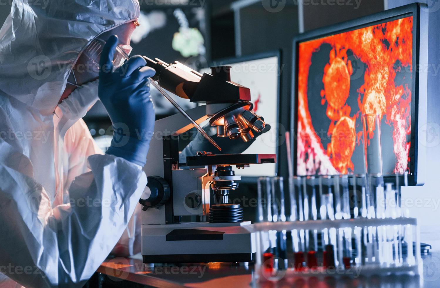 cientista em uniforme protetor branco perto de monitores com dados trabalha com coronavírus e tubos de sangue em laboratório foto