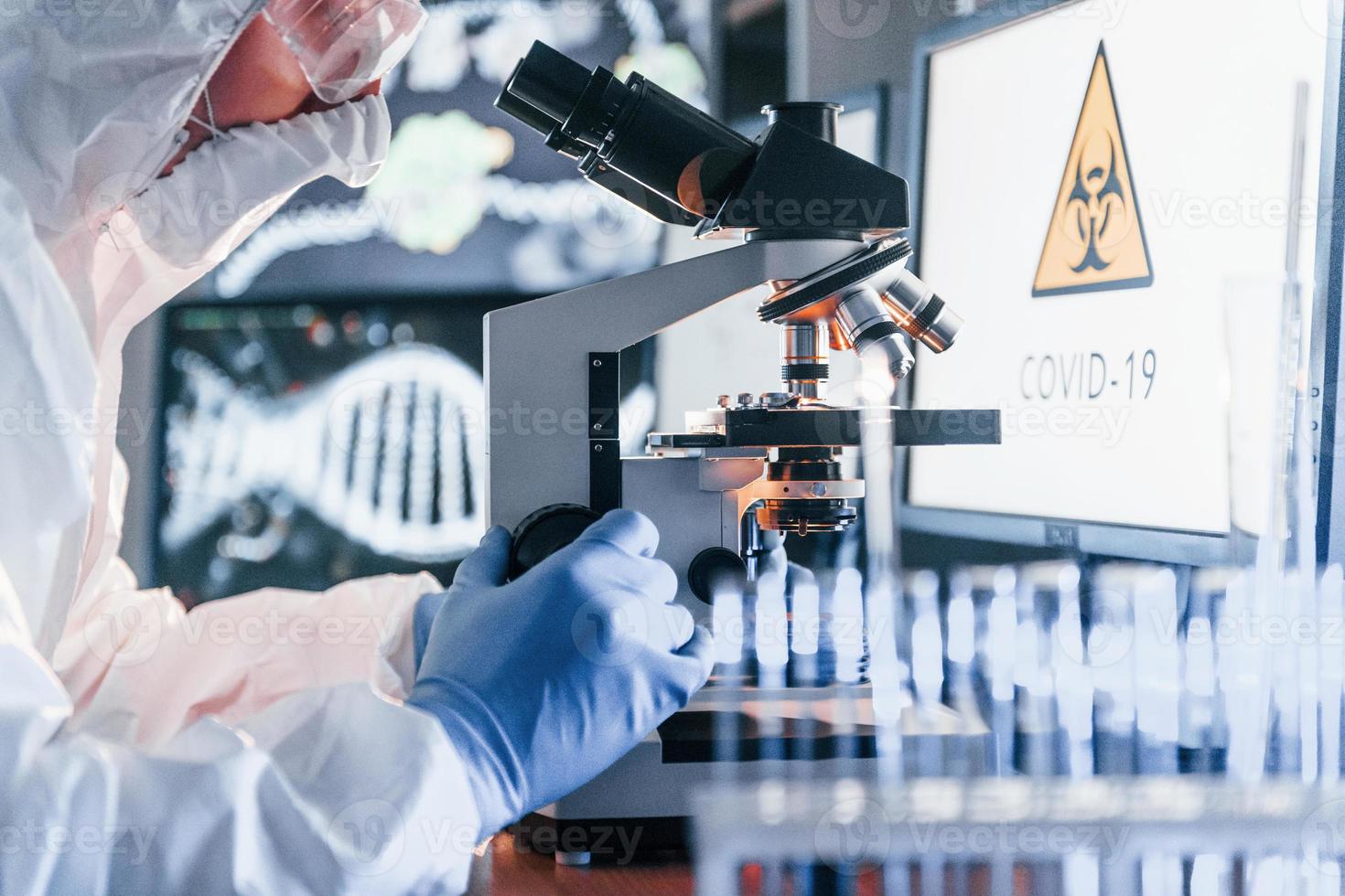 monitores com informações sobre a mesa. cientista em uniforme protetor branco trabalha com coronavírus e tubos de sangue em laboratório foto