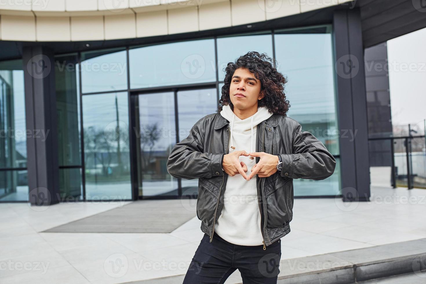 jovem bonito com cabelo preto encaracolado posando para a câmera na rua contra a construção foto