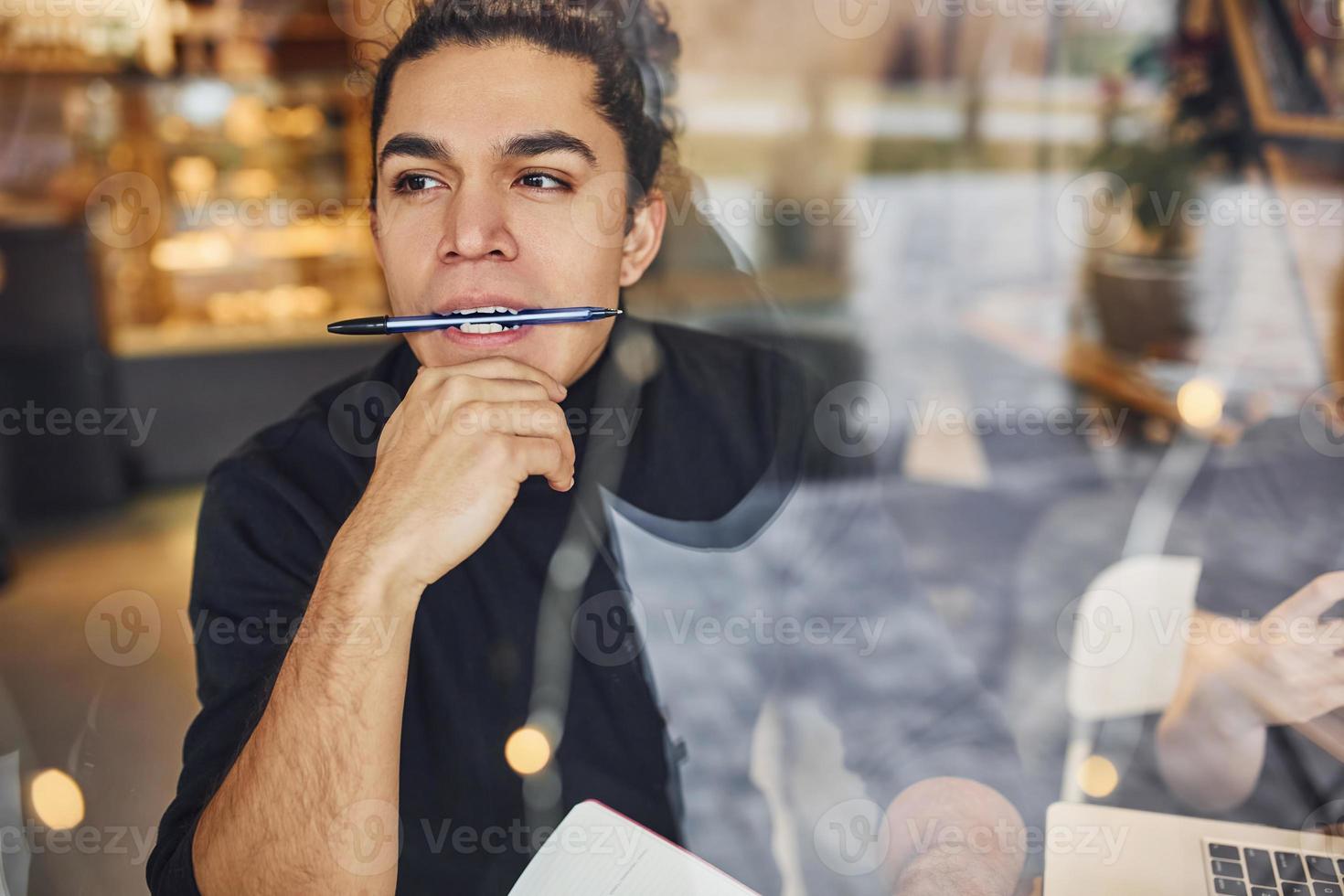 jovem escritor masculino com cabelo preto encaracolado sentado dentro de casa no café com caneta e bloco de notas. ver através do vidro foto