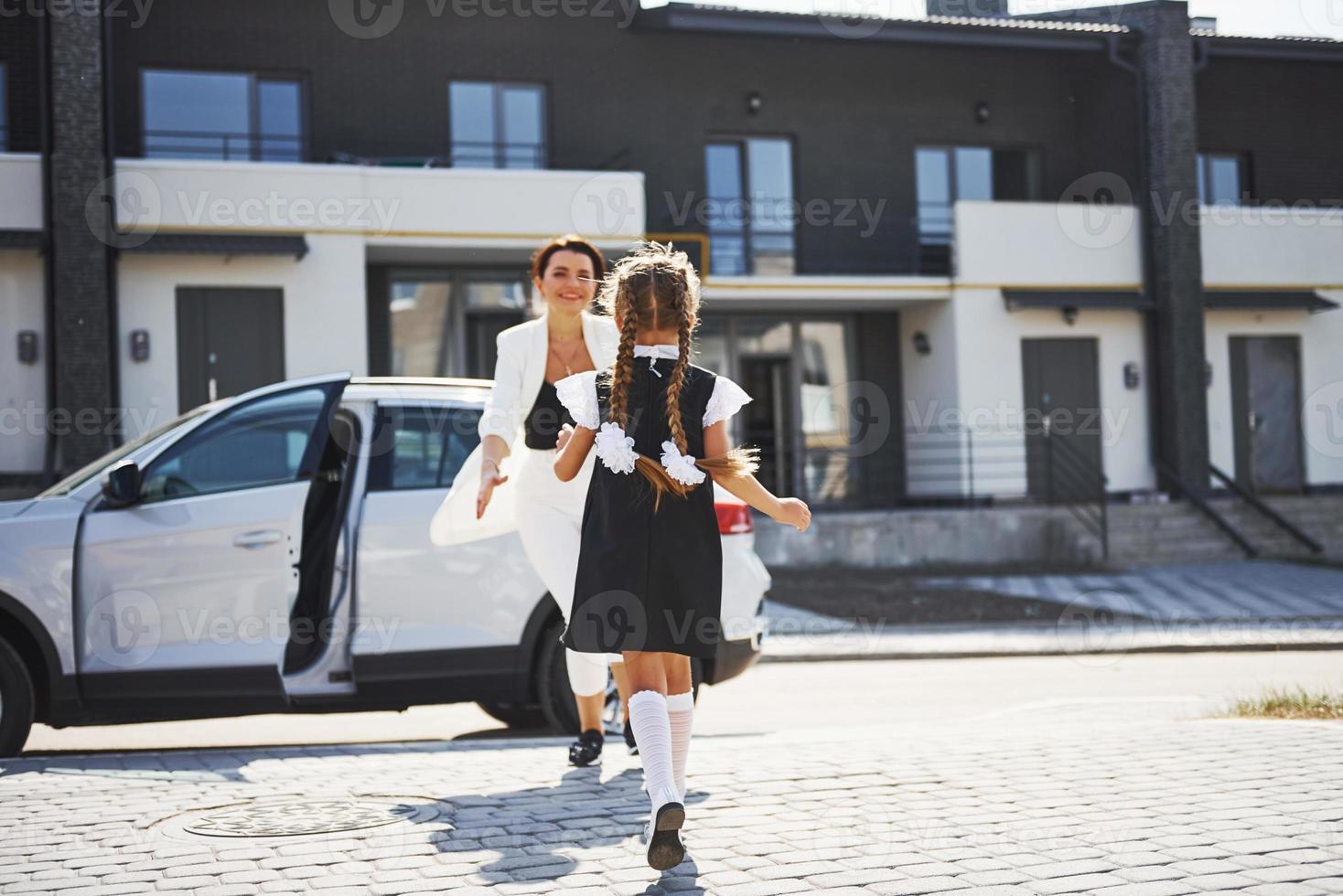 mãe com filha em uniforme escolar ao ar livre perto de carro branco foto