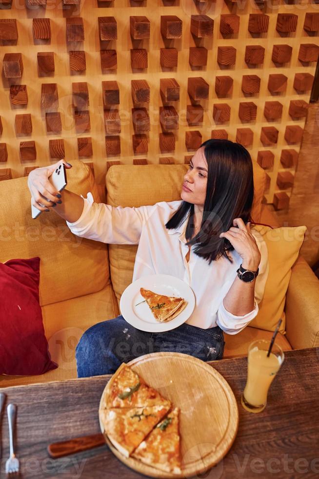 jovem morena sentada dentro de casa com pizza e smartphone na mão foto