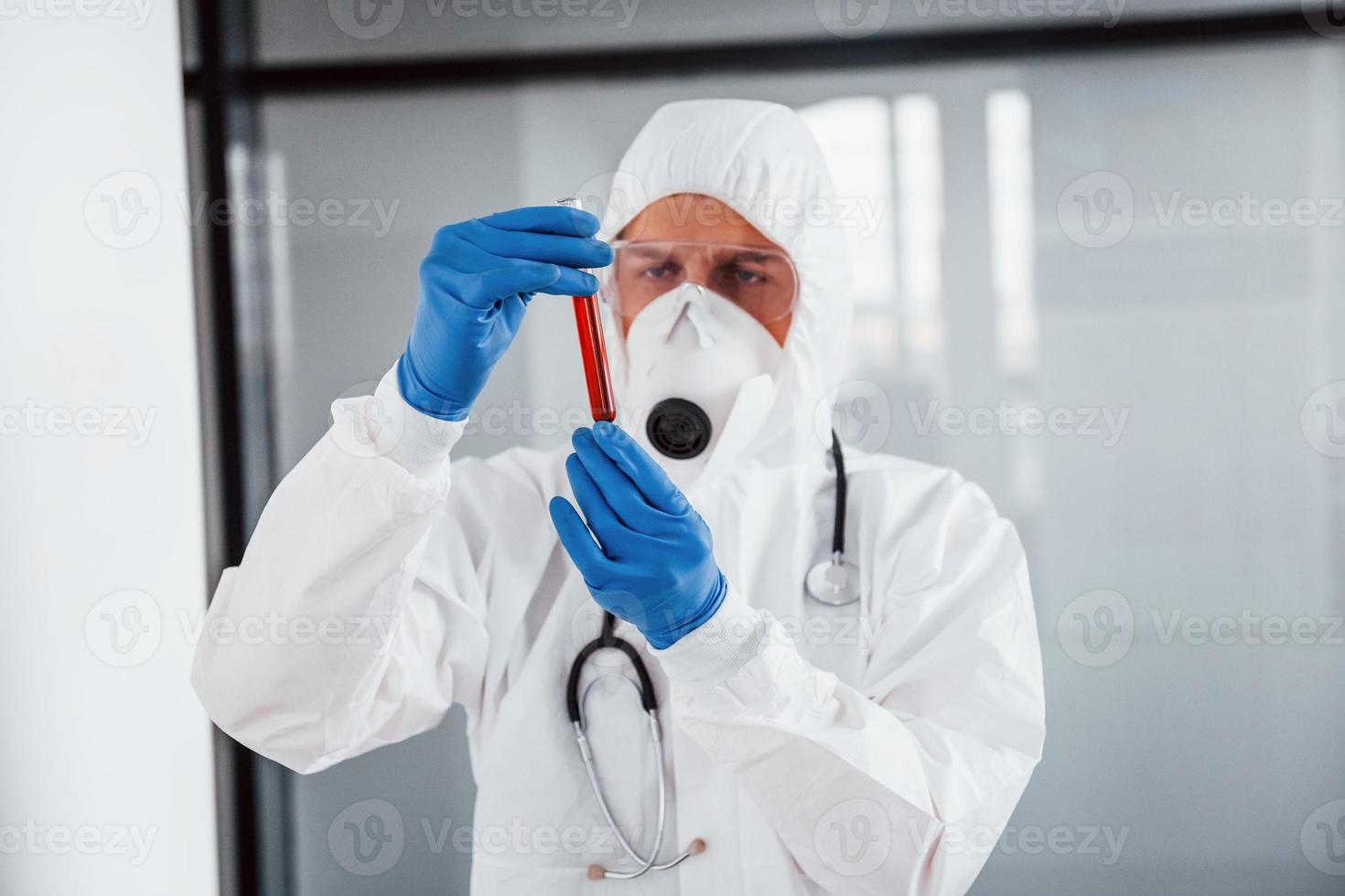 cientista médico masculino em jaleco, óculos de proteção e máscara segura tubo de ensaio com sangue foto