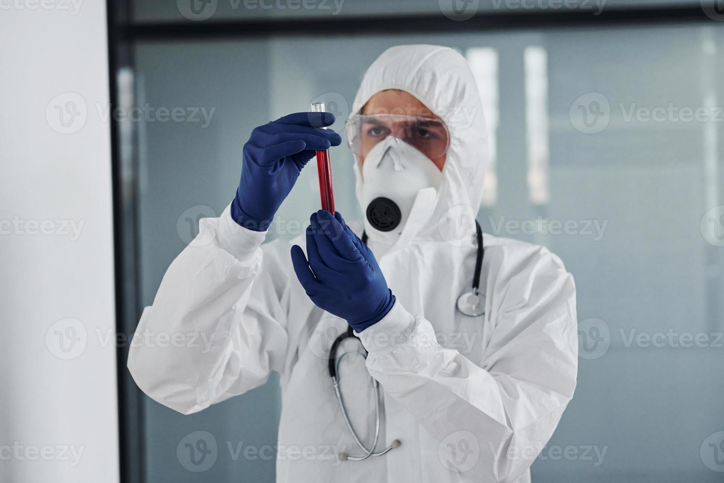 cientista médico masculino em jaleco, óculos de proteção e máscara segura tubo de ensaio com sangue foto
