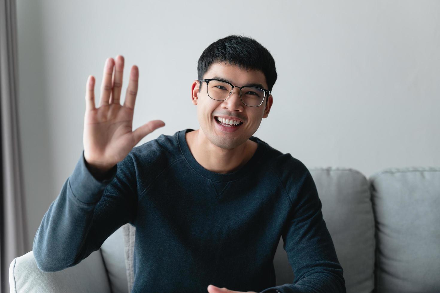 retrato de jovem asiático usando óculos grandes sorrindo e olhando para a câmera com sentimento feliz e positivo na sala de estar em casa, reunião, videoconferência. foto