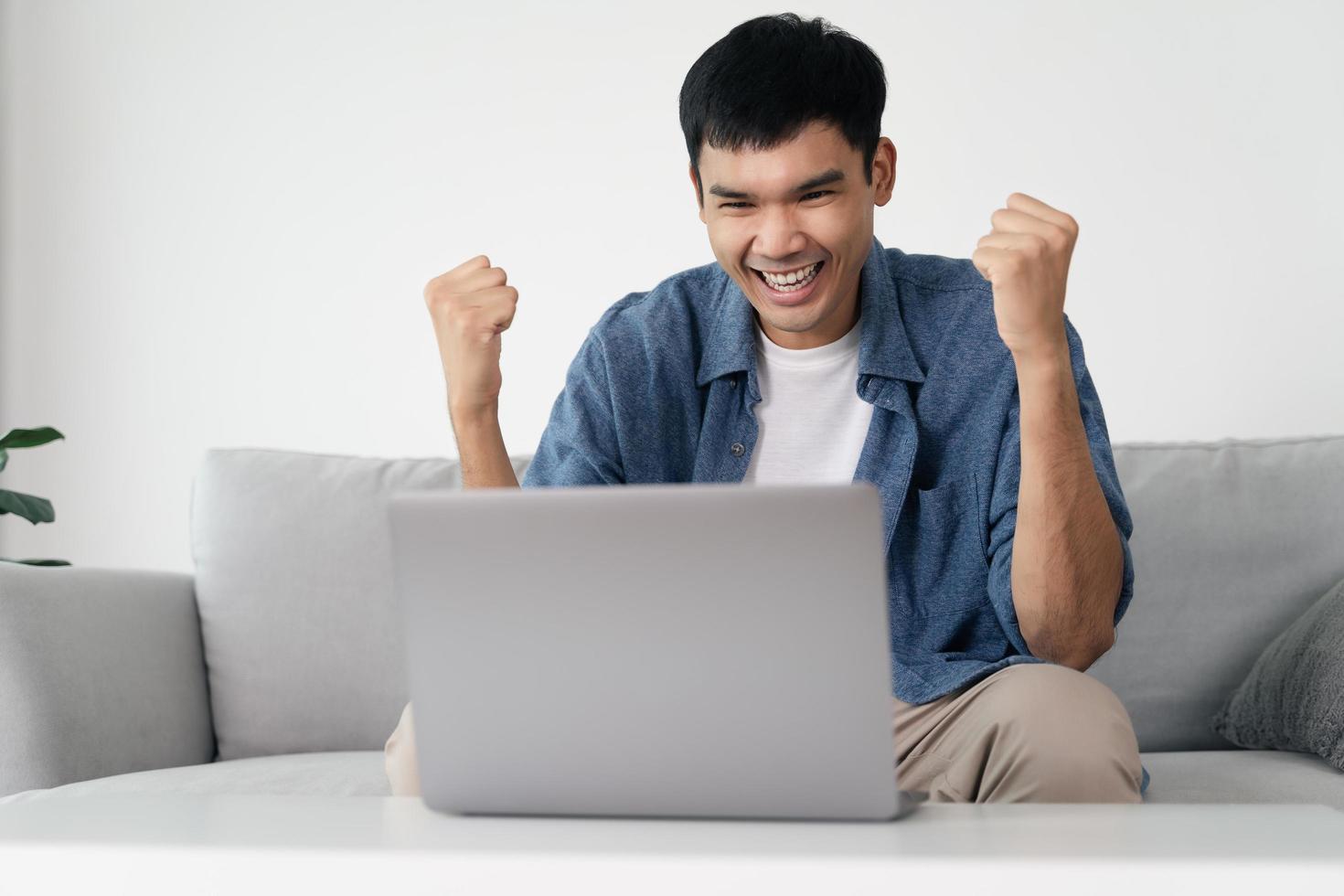 feliz homem asiático animado olhando para laptop, homem recebendo boas notícias usando o computador em casa. foto