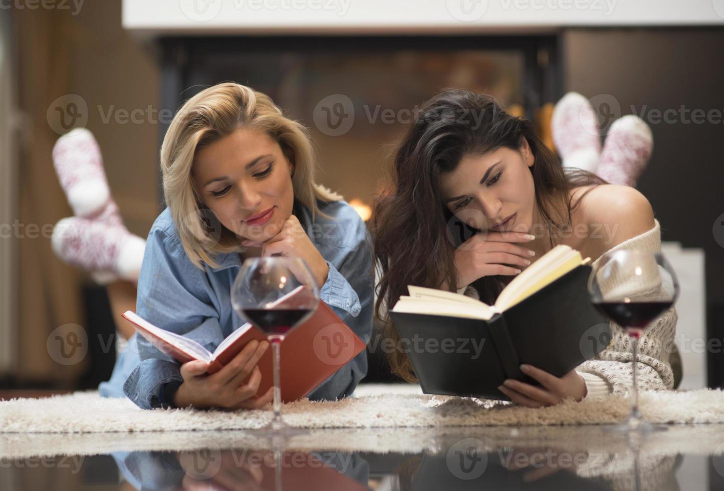 duas amigas lendo um livro e bebendo vinho tinto perto de uma lareira. conceito de estilo de vida. foto