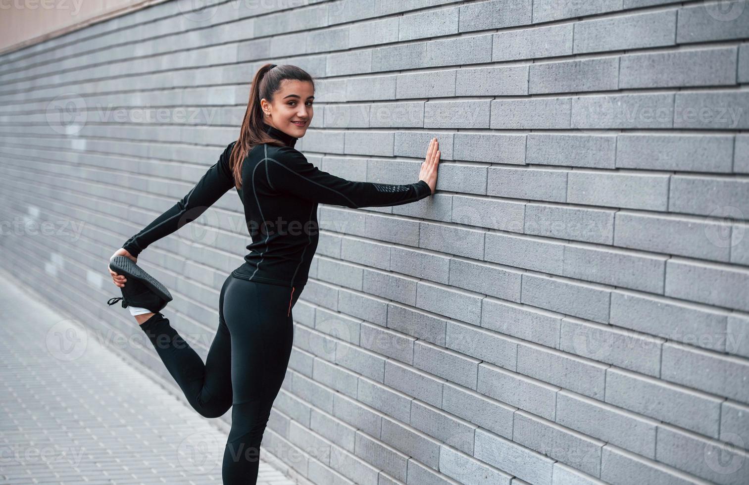 jovem esportiva em roupas esportivas pretas ao ar livre fazendo alongamento perto da parede cinza foto