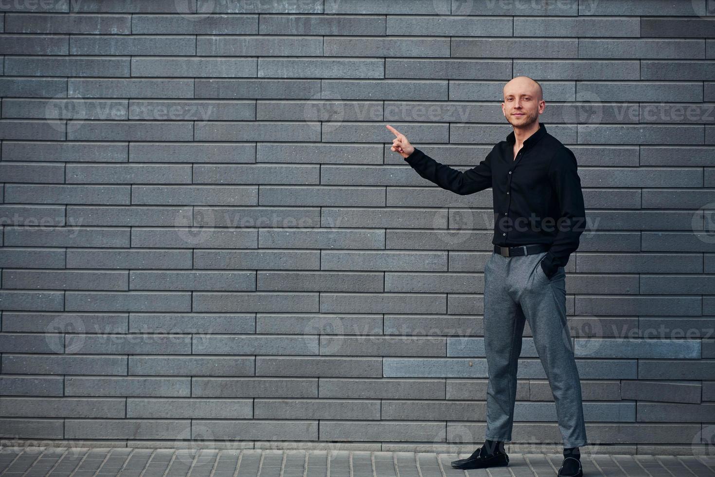 homem elegante elegante em pé ao ar livre contra a parede cinza e mostra à esquerda pelo dedo foto