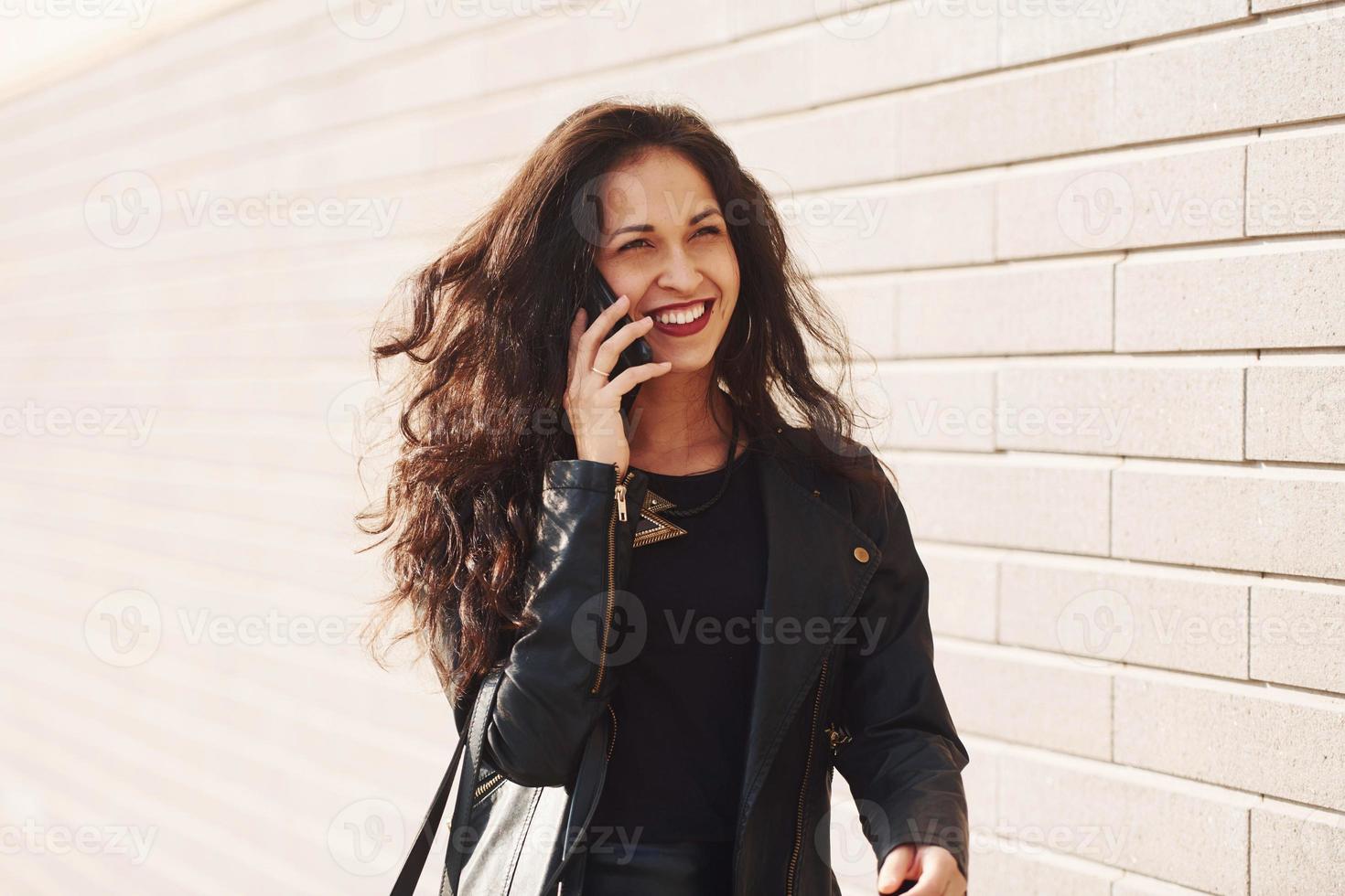 retrato de morena alegre com cabelos cacheados e roupas pretas encostado na parede e fala por telefone foto