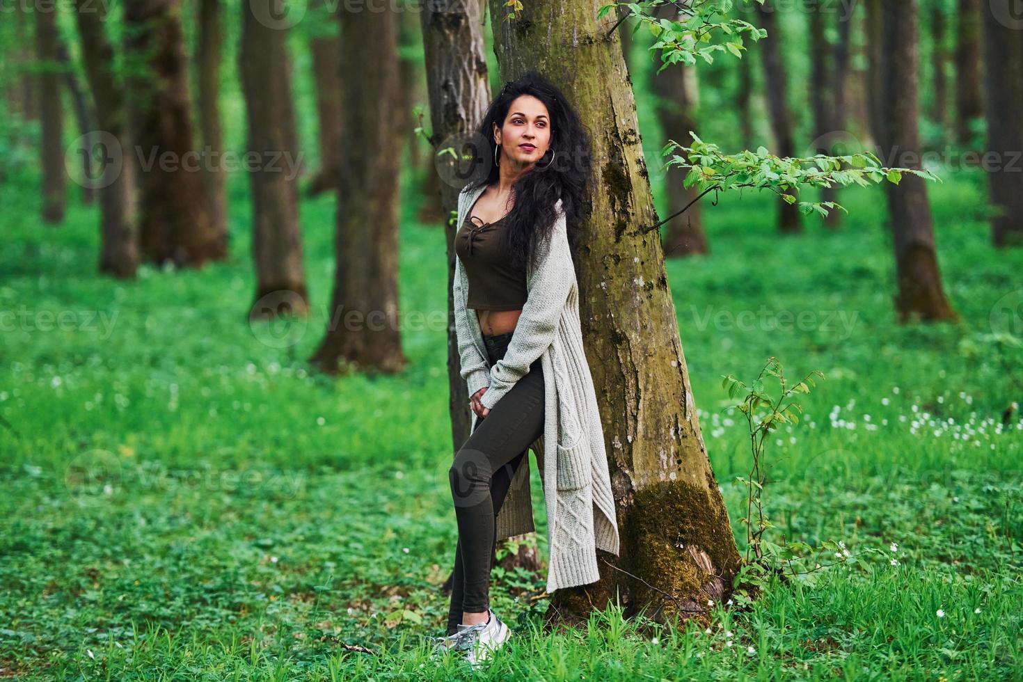 bela morena em jeans preto em pé na floresta perto da árvore foto