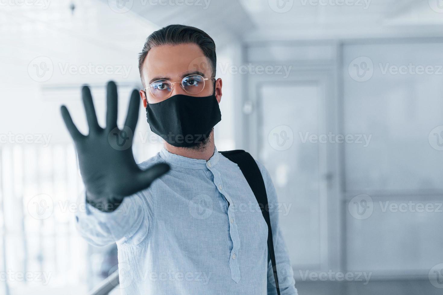 mostrando o gesto de parada com a mão. em máscara protetora e luvas. jovem bonito em roupas formais dentro de casa no escritório durante o dia foto