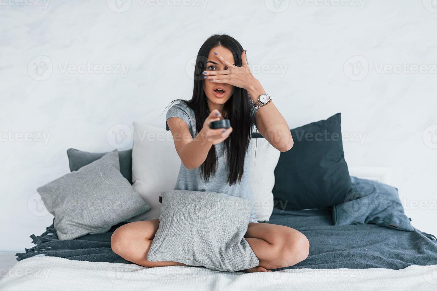segurando o controle remoto da tv e assistindo filme. jovem mulher bonita em roupas casuais sentada em casa sozinha foto