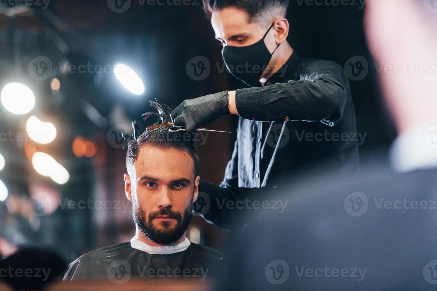 vista frontal do jovem barbudo sentado e cortando o cabelo na barbearia por cara com máscara protetora preta foto